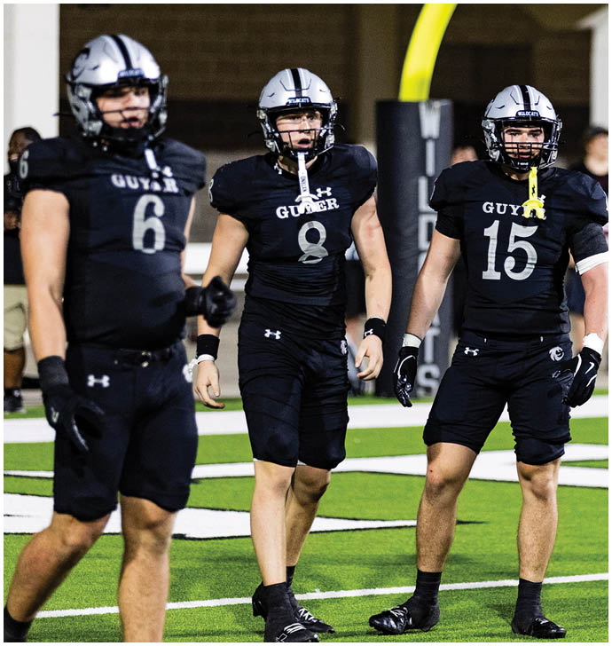 Three people wearing black football uniforms and helmets stand on a field near a yellow goalpost. Description generated by AI