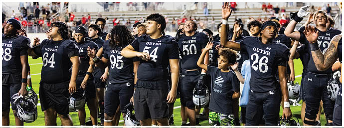 A group of people wearing matching sports jerseys are gathered on a field with empty stadium seats in the background, some raising their hands and appearing engaged or celebratory. Description generated by AI