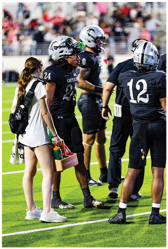 Two football players in helmets and uniforms are on the field with a coach wearing a headset, with blurred spectators in the background. Description generated by AI