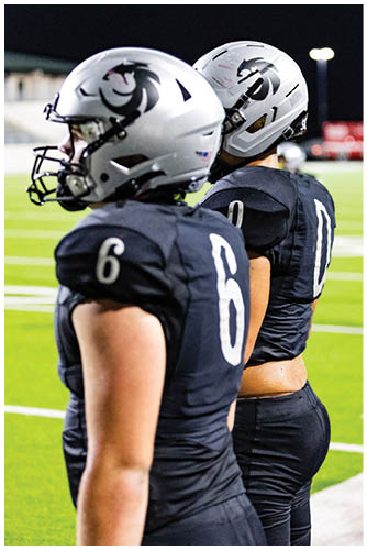 Two people wearing white football helmets with a black lion logo are shown against a dark background. Description generated by AI