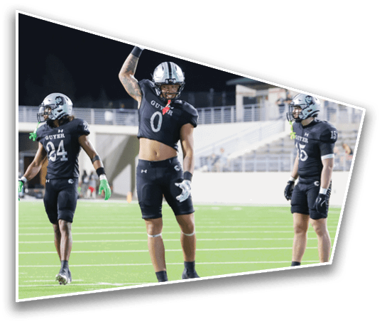 Three people in black football uniforms with helmets stand on a green field, with one person raising an arm in celebration. Description generated by AI