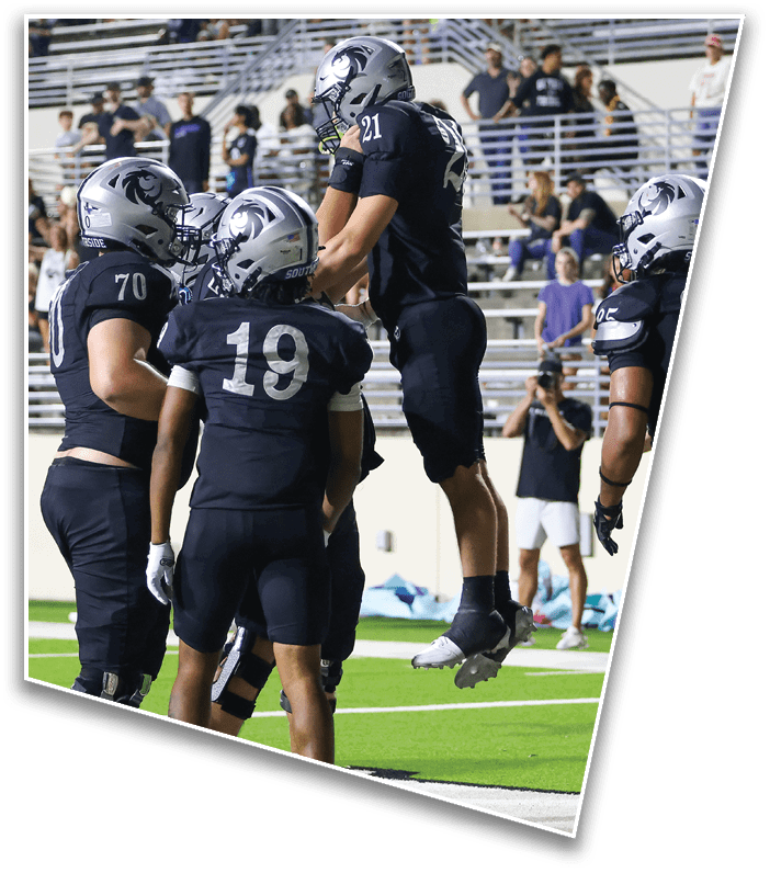 Several football players in dark uniforms and helmets are on a field with one player jumping while others stand nearby and spectators watch from the bleachers. Description generated by AI