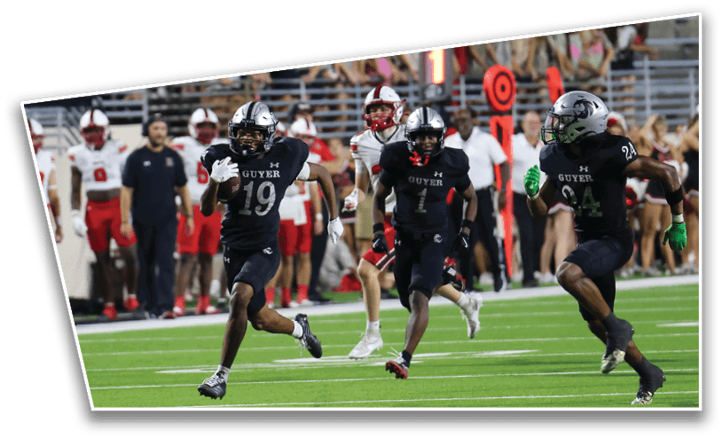 Three people in black football uniforms with helmets are running on a green field while people in red and white uniforms stand in the background. Description generated by AI