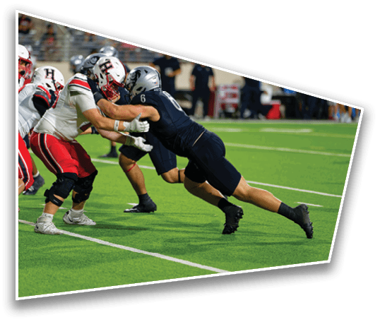Two football players in helmets and uniforms are engaged in a physical block on a green field during a game. Description generated by AI