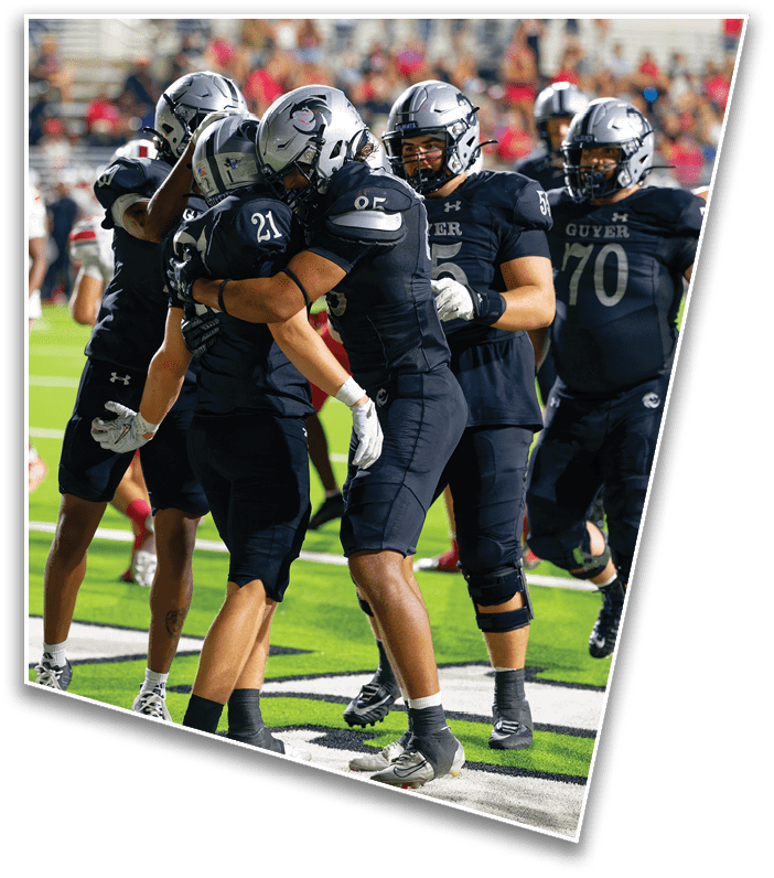 Four football players in black uniforms and helmets are celebrating on the field while other players in white and red uniforms stand in the background. Description generated by AI