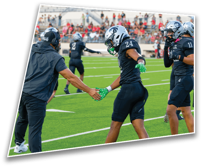People in football uniforms are on a field with one player shaking hands with a person on the sideline while others stand nearby. Description generated by AI