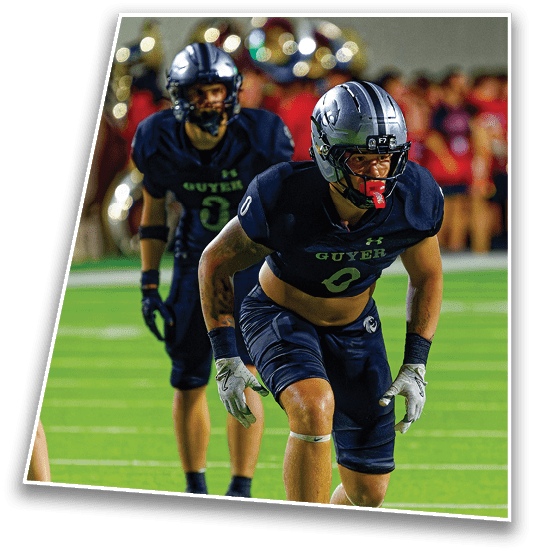 Two people in dark football uniforms and helmets are on a green field with one person in a ready stance and the other standing behind. Description generated by AI