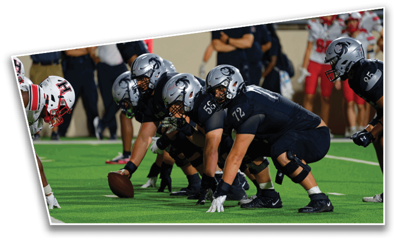 A group of football players in dark uniforms and helmets are lined up on the field in a ready stance facing opponents in white and red uniforms. Description generated by AI