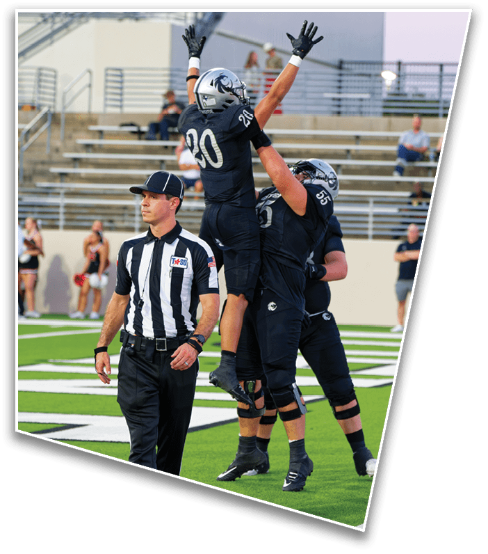 A person wearing a black football uniform with the number 20 and a silver helmet raises both arms in celebration on a football field with spectators in the background. Description generated by AI