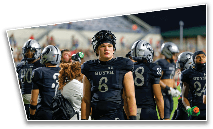 A group of people wearing black football uniforms with helmets stand on a field, with one person in the center looking forward and others engaged in various activities. Description generated by AI