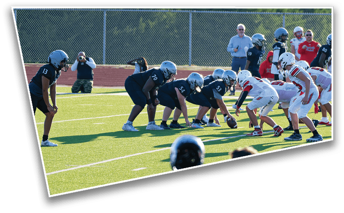 Two teams of people in football uniforms are lined up on a field preparing for a play while others and coaches stand on the sidelines. Description generated by AI