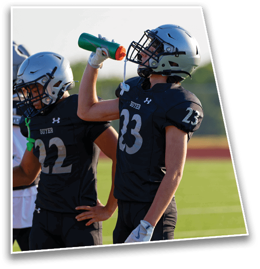People wearing football uniforms and helmets are standing together on a field, with one person holding a water bottle above another's shoulder. Description generated by AI
