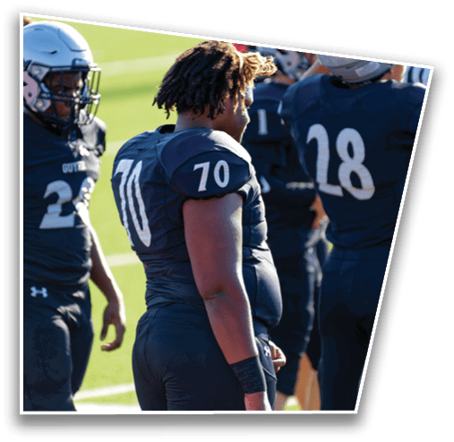 People wearing dark football uniforms with numbers stand on a grassy field, some holding helmets. Description generated by AI