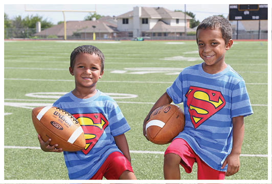 Two children wearing matching blue striped shirts with a Superman logo and red shorts are smiling and holding footballs on a sports field. Description generated by AI