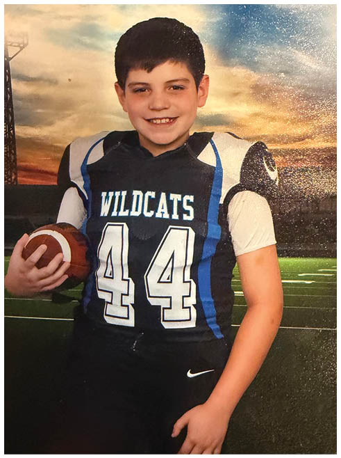 A child wearing a Wildcats football uniform with the number 44 is holding a football and smiling on a football field at sunset. Description generated by AI