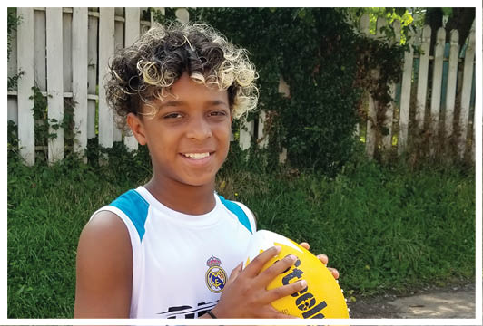 A child with curly hair wearing a sports jersey is smiling and holding a yellow football outdoors near a white fence and greenery. Description generated by AI
