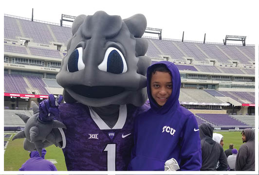 A person wearing a purple hoodie stands next to a large mascot in a purple sports jersey inside a stadium. Description generated by AI