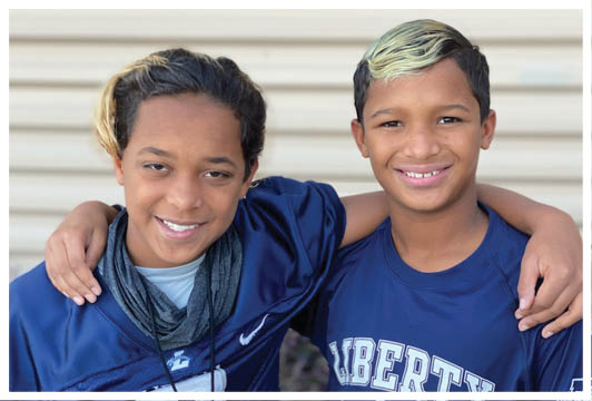 Two children with short hair and casual clothing smile with their arms around each other in front of a light-colored wall. Description generated by AI
