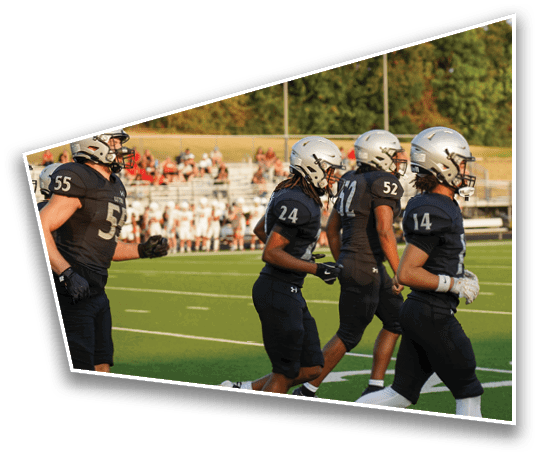 Four people wearing black football uniforms and silver helmets are walking on a football field with a blurred crowd in the background. Description generated by AI