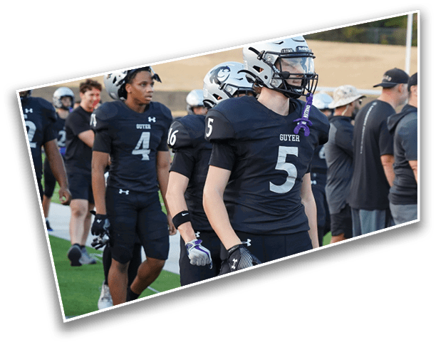 People wearing black football uniforms with numbers and helmets are standing on a field, with some wearing gloves and mouthguards. Description generated by AI