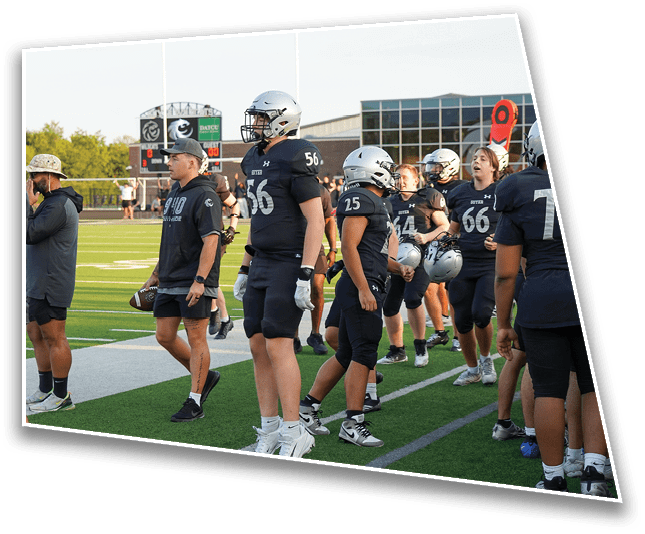 People in football uniforms and coaches stand on the sidelines of a football field during a game or practice. Description generated by AI