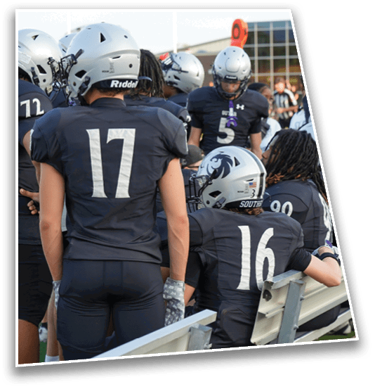 People wearing dark sports uniforms with numbers are sitting and standing near a bench on a sports field. Description generated by AI