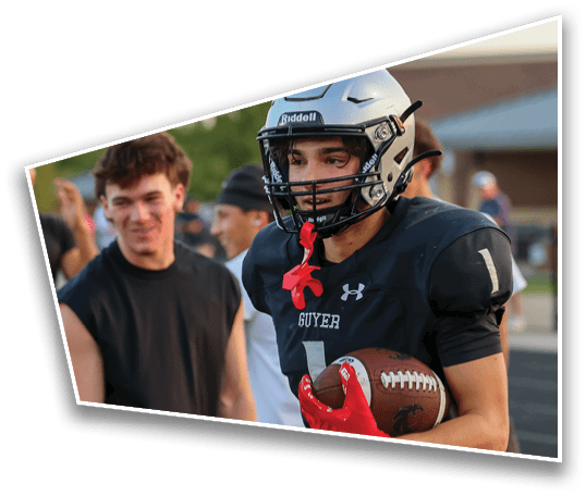 A person wearing a football helmet and black jersey with the word “GUYER" is holding a football on a sports field. Description generated by AI