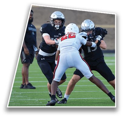 Three people in football uniforms are engaged in a play on a green field with one person in white blocking two people in black, while another person in black stands nearby. Description generated by AI