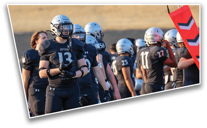 A group of people wearing black football uniforms and silver helmets stand on a field near an orange marker with black arrows. Description generated by AI