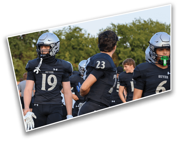 People wearing football uniforms and helmets are standing outdoors on a field with trees in the background. Description generated by AI