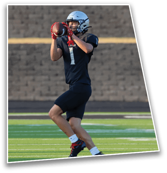 A person wearing a helmet and sports uniform is catching a football on a green field with a brick wall in the background. Description generated by AI