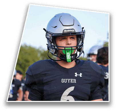 A person wearing a silver football helmet and a black jersey with the word “GUYER" stands outdoors with blurred people and trees in the background. Description generated by AI