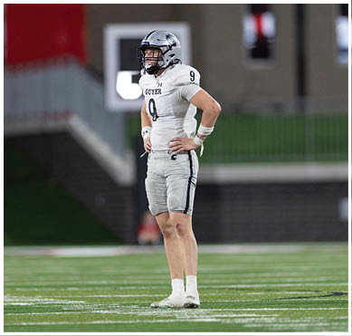 A person wearing a football uniform and helmet stands on a field with hands on hips, looking to the side. Description generated by AI