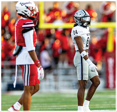 Two people in football uniforms stand on a field with a blurred marching band in the background. Description generated by AI