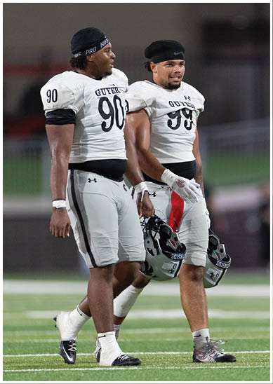 Two adults wearing football uniforms walk side by side on a field holding helmets in their hands. Description generated by AI