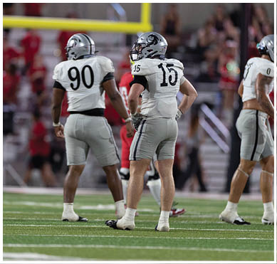 Three people in white and gray football uniforms stand on a field with a blurred crowd in the background. Description generated by AI