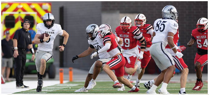 A person in a white sports uniform is running with a football while several people in red and white uniforms chase on a green field with officials and spectators in the background. Description generated by AI