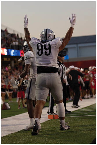 A person wearing a white football uniform with the number 99 raises both arms in celebration on a football field with other players and officials in the background. Description generated by AI
