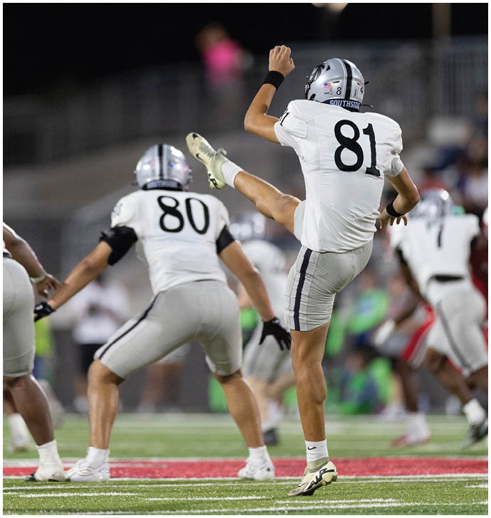 Two people in white football uniforms with helmets are on a field, with one person kicking a ball and the other preparing to catch or block. Description generated by AI