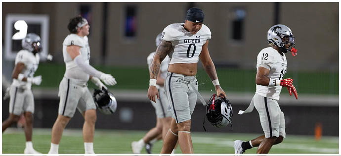 Four people in football uniforms walk on a field with one holding a helmet and another wearing a helmet with a mouthguard. Description generated by AI