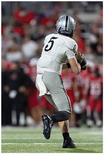 A person wearing gray athletic pants and black cleats is running on a football field with a crowd of people in red uniforms blurred in the background. Description generated by AI