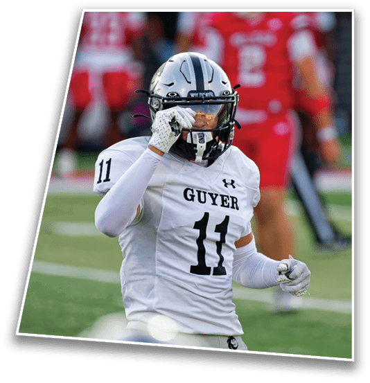 A person wearing a white football uniform with the number 11 and the word Guyer on the jersey is adjusting their helmet on a football field with players in red uniforms blurred in the background. Description generated by AI