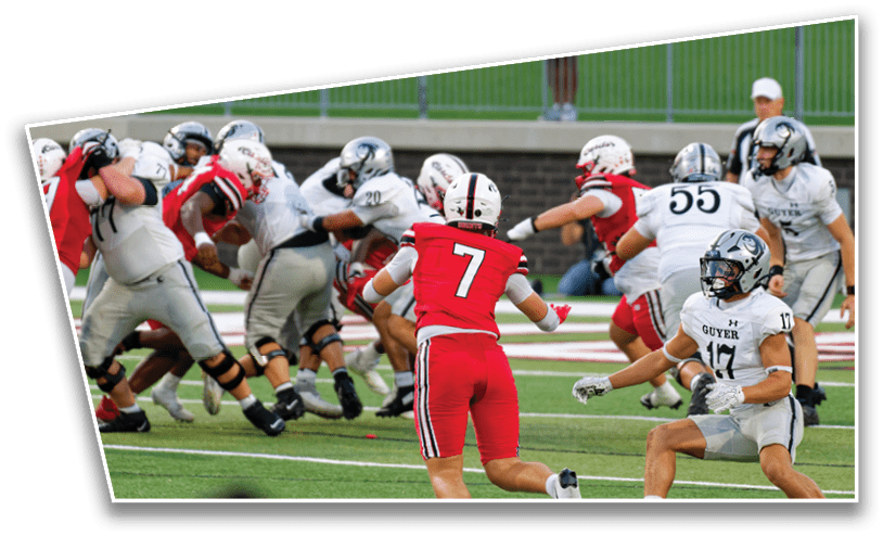 People in red and white sports uniforms are engaged in a football game on a green field with a fence and spectators in the background. Description generated by AI