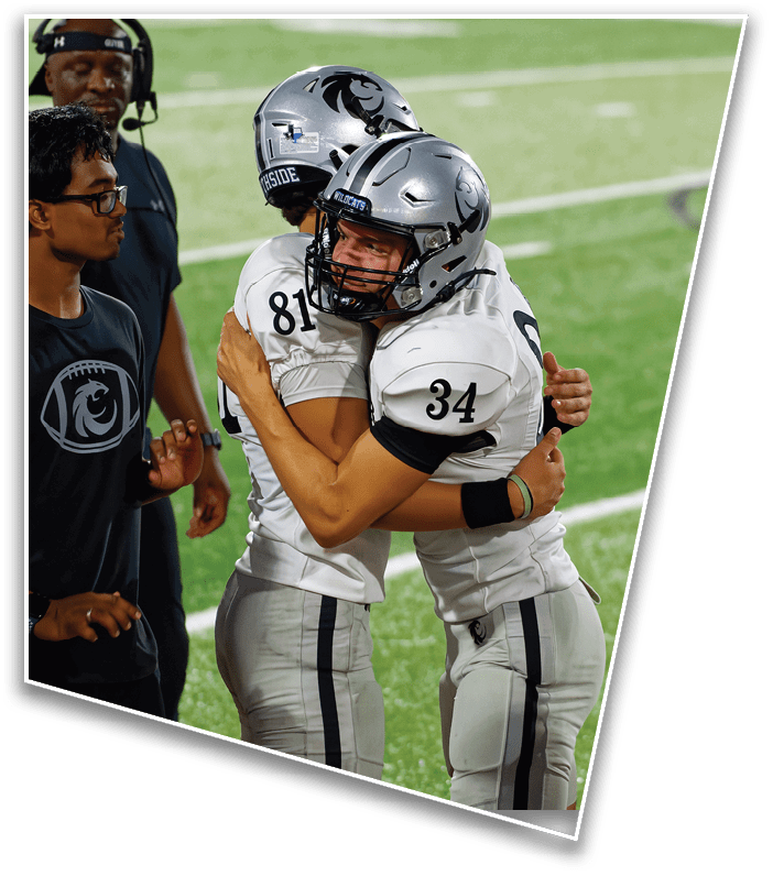 Two people wearing football uniforms and helmets are embracing on a football field. Description generated by AI