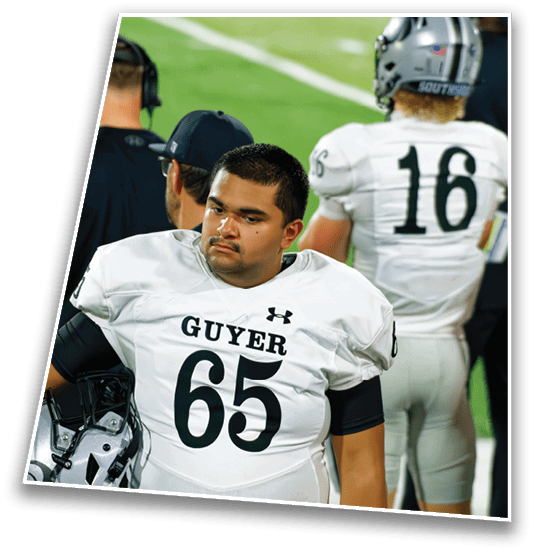 A person wearing a white football uniform with the number 65 and the word Guyer stands on a field holding a helmet while another person in a white uniform with the number 16 stands nearby. Description generated by AI