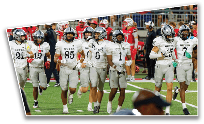 A group of football players in white and gray uniforms with helmets are walking together on a field with players in red uniforms visible in the background. Description generated by AI