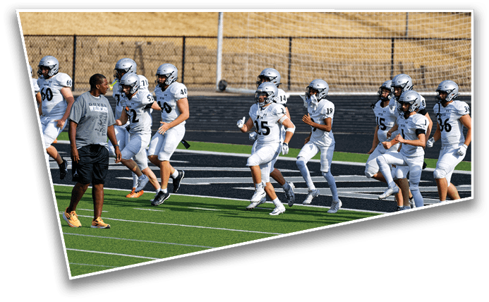 A group of people in white football uniforms and helmets are running on a field alongside a person in a gray shirt and black shorts. Description generated by AI