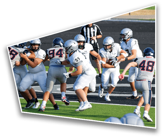 A group of people wearing football helmets and uniforms are actively engaged in a play on a football field with a referee observing in the background. Description generated by AI
