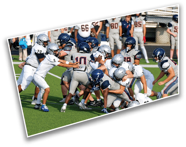 A group of people wearing football helmets and uniforms are engaged in a play on a green field with others standing in the background. Description generated by AI