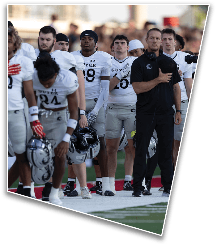 A group of people in white football uniforms stand in a line on a field with one person in black clothing holding a hand over their chest. Description generated by AI